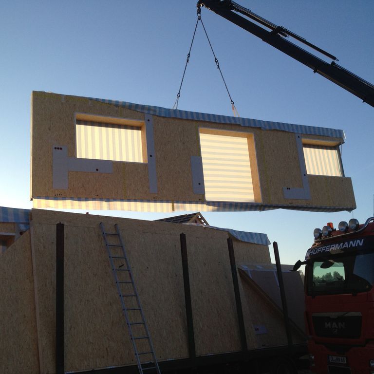 Foto eines Haus-Fertigbauteils schwebend an einem Kran. Es sind Ausschnitte für eine Doppeltür in der Mitte und je ein Fenster rechts und links erkennbar. Das Fertigbauteil ist aus Holz.